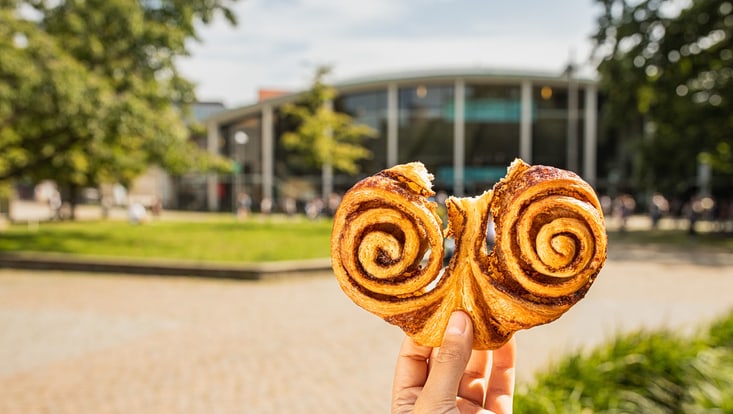 Franzbrötchen in der Hand vorm Hintergrund des Audimax